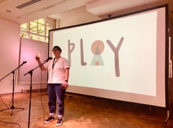Nic Golightly on stage in front of a screen showing the word 'play'